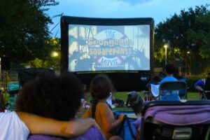Families watch "The SpongeBob SquarePants Movie" on a large outdoor screen at dusk.