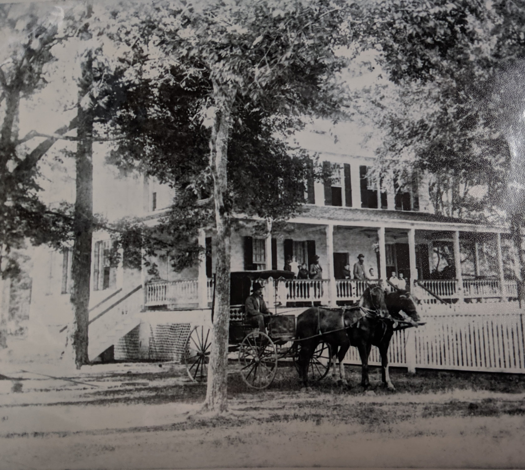 A horse-drawn carriage is parked in front of a large, two-story house with people on the porch.