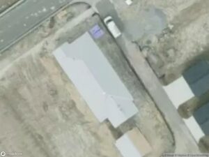 Aerial view of a gray building near a road, with a white vehicle parked nearby. Surrounding area is mostly barren.