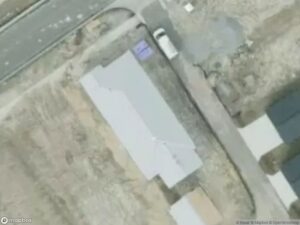 Aerial view of a residential area with a house, road, and parked vehicle. Surrounding land appears mostly bare.