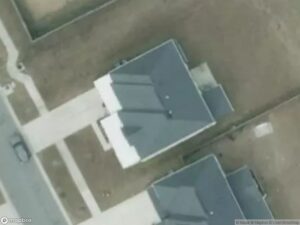 Aerial view of two houses with gray roofs and a driveway, surrounded by bare land.