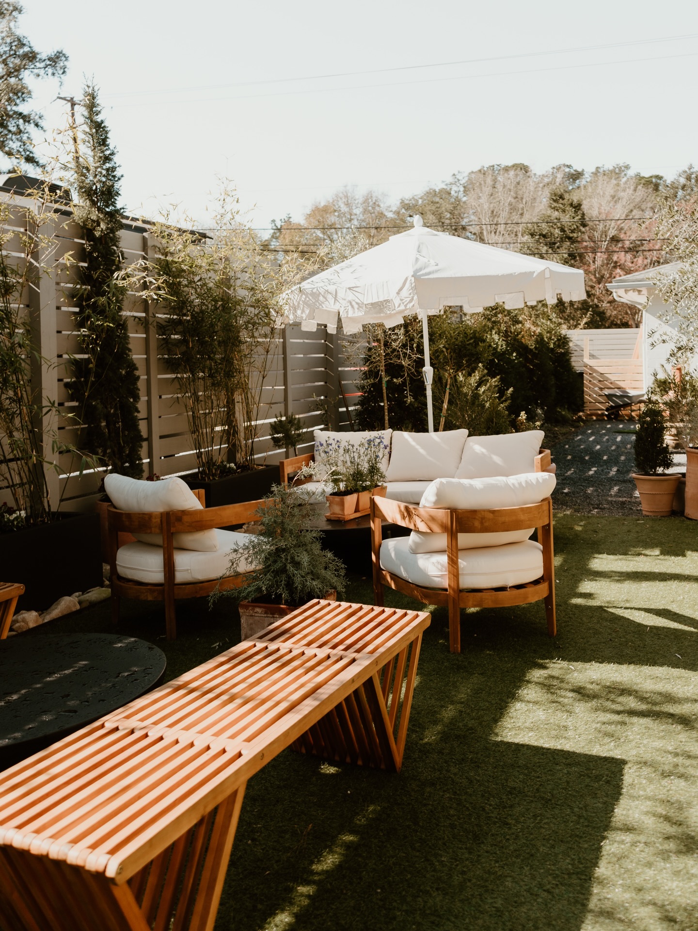 A cozy outdoor seating area with wooden chairs, a table, and a large umbrella surrounded by greenery.