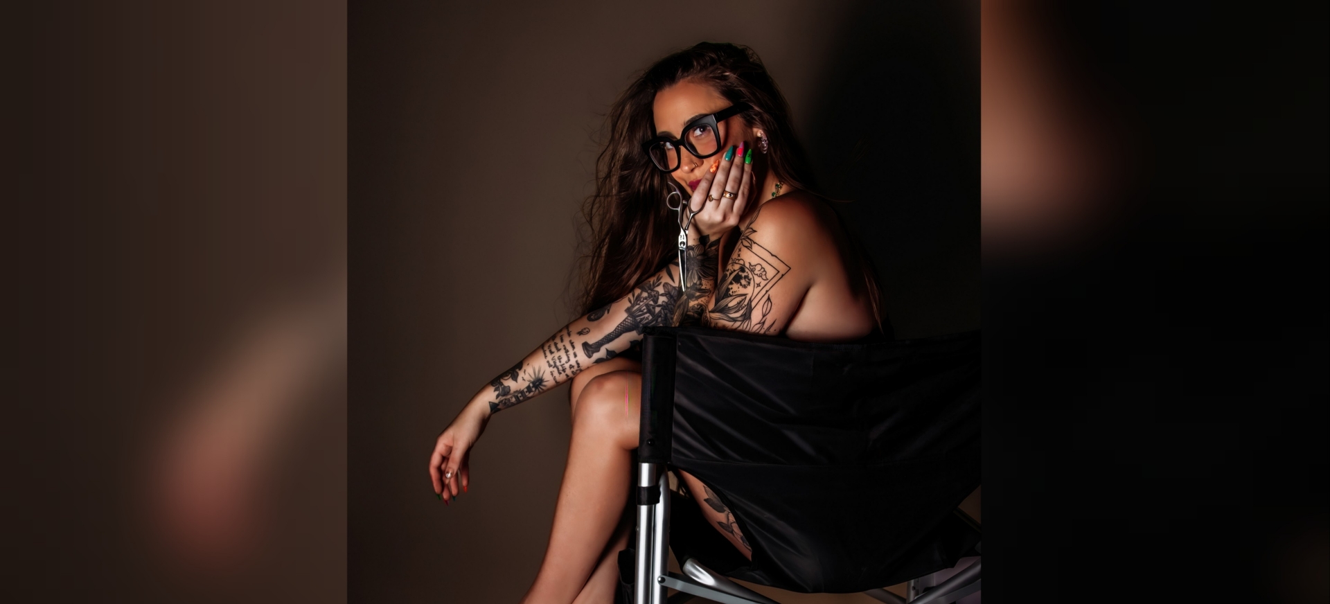 A person with tattoos sits in a chair, resting their chin on their hand, wearing glasses and colorful nails.