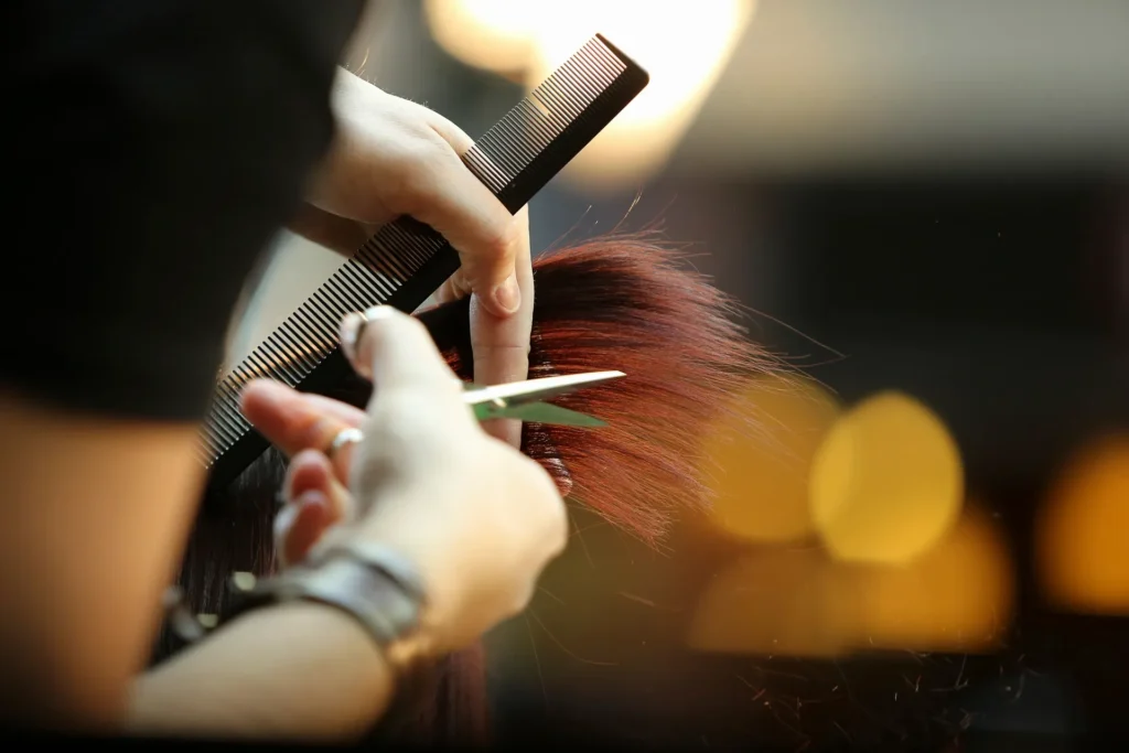 A stylist uses scissors and a comb to cut red hair. Blurred lights in the background.