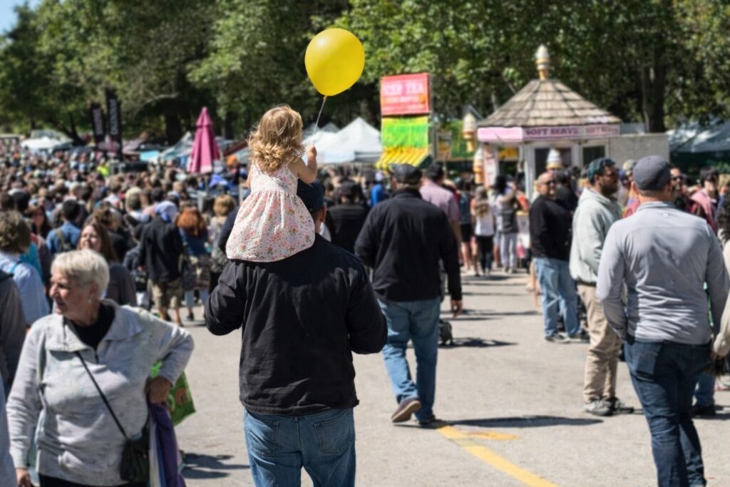 A child with a yellow balloon sits on a man's shoulders in a crowded festival setting.