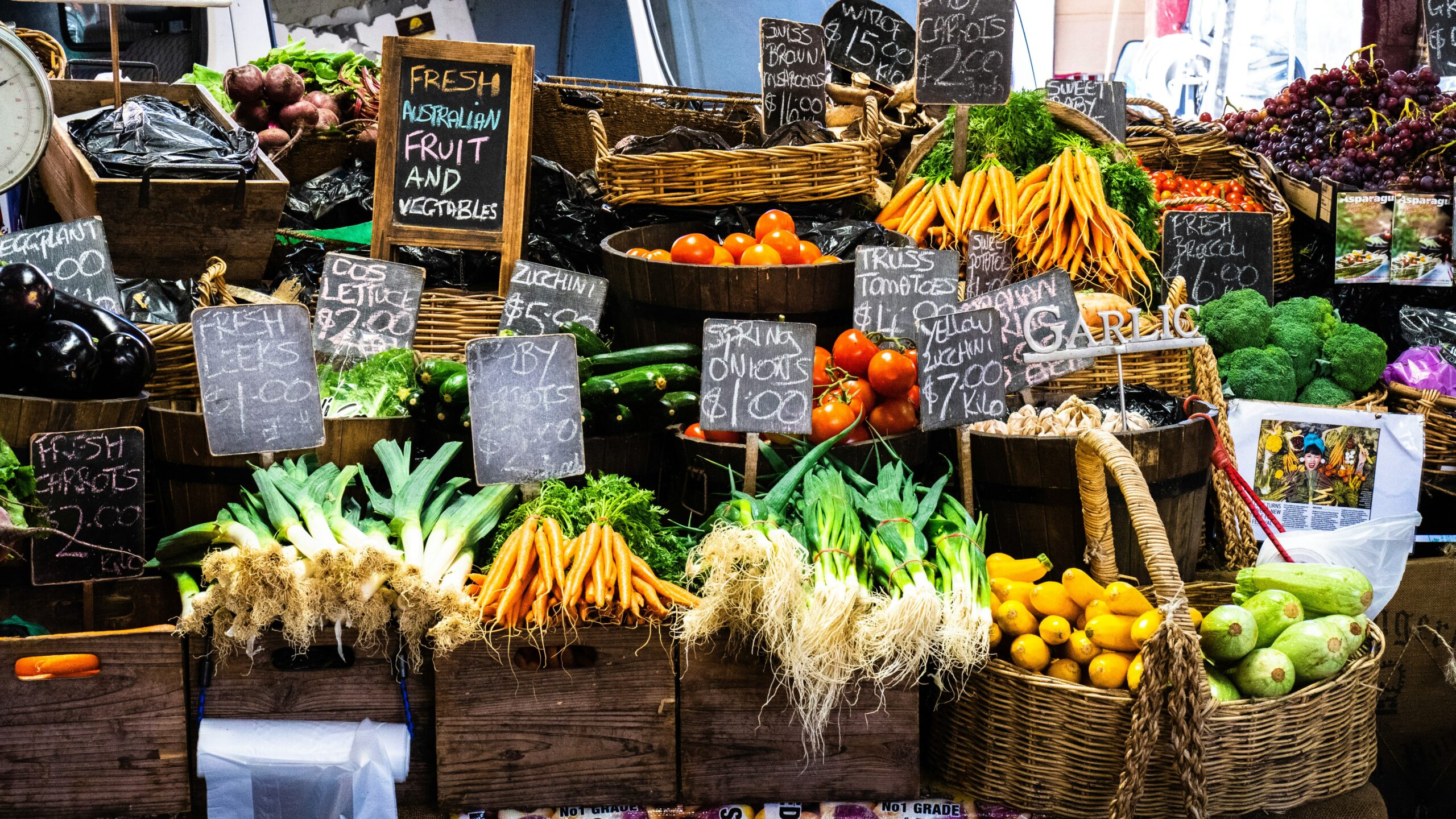 A vibrant market display of fresh fruits and vegetables with price tags on wooden crates and baskets.