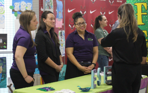 Four women in purple shirts engage in conversation at a table with various items displayed.