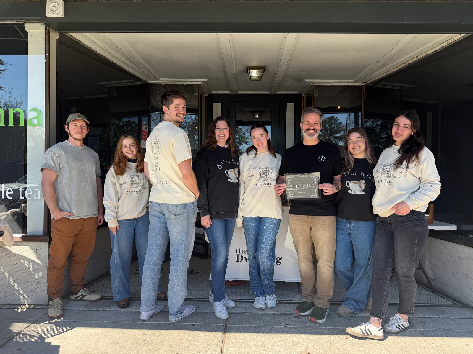 A group of eight people stands outside a café, smiling and holding a certificate.