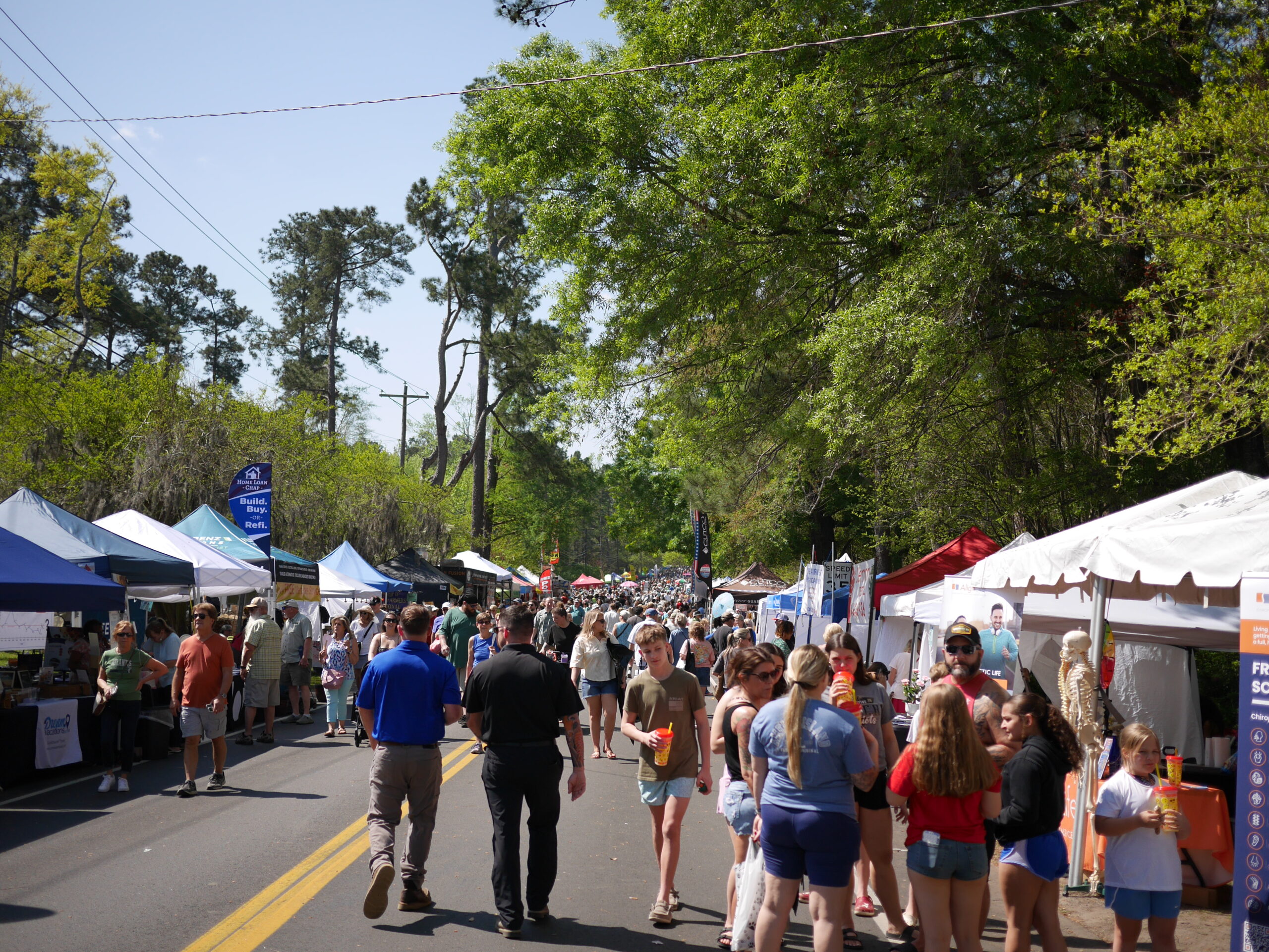 Crowd gathers at a street fair with tents, vendors, and people enjoying the sunny day.