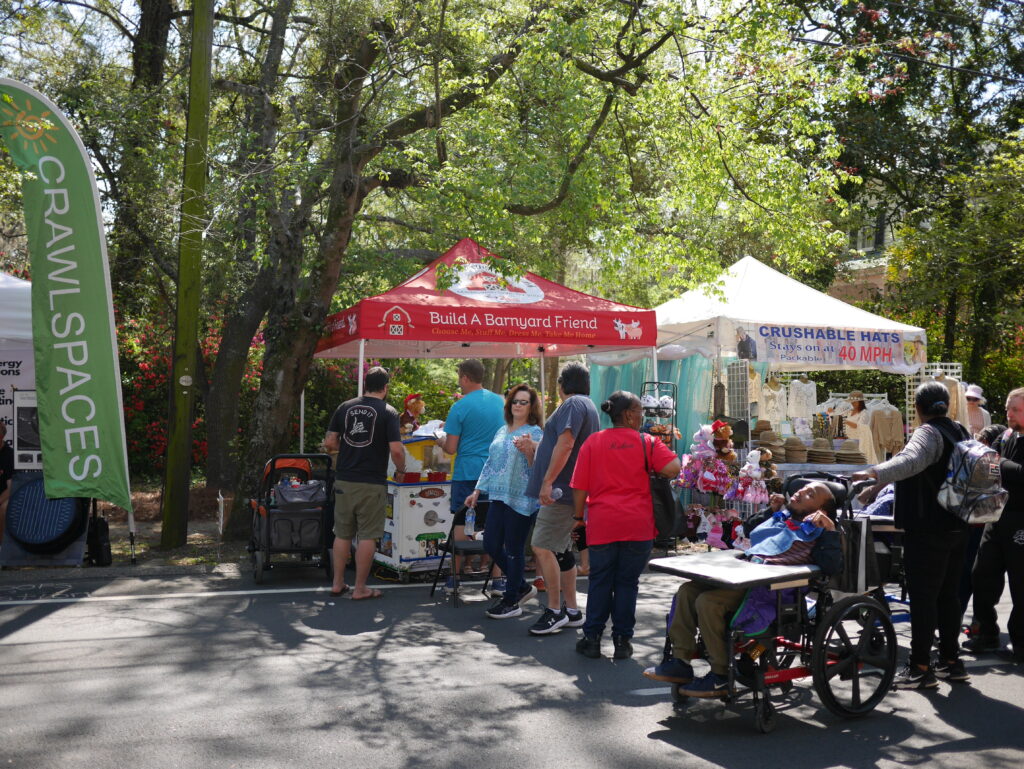 People gather at vendor tents, browsing items and enjoying the outdoor market atmosphere. A child in a wheelchair is present.