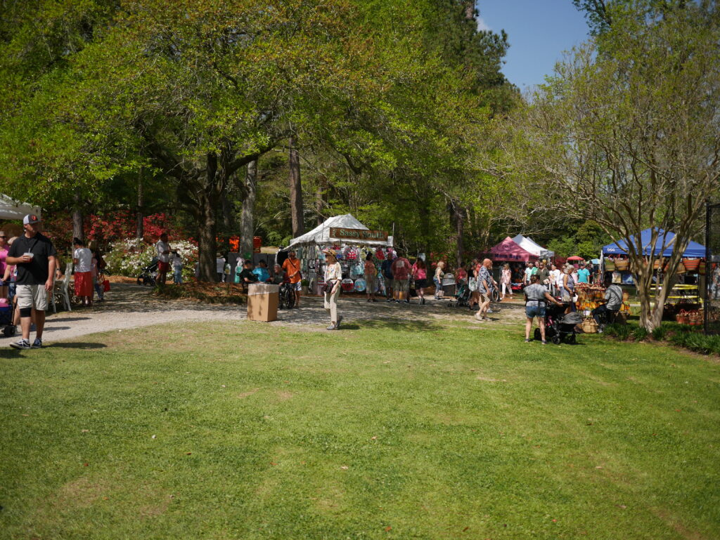 Crowd gathers at a lively outdoor market with tents, trees, and colorful flowers in the background.