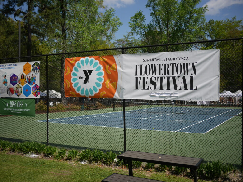 Banners for the Flowertown Festival and YMCA displayed near a tennis court. Benches and tents are visible in the background.