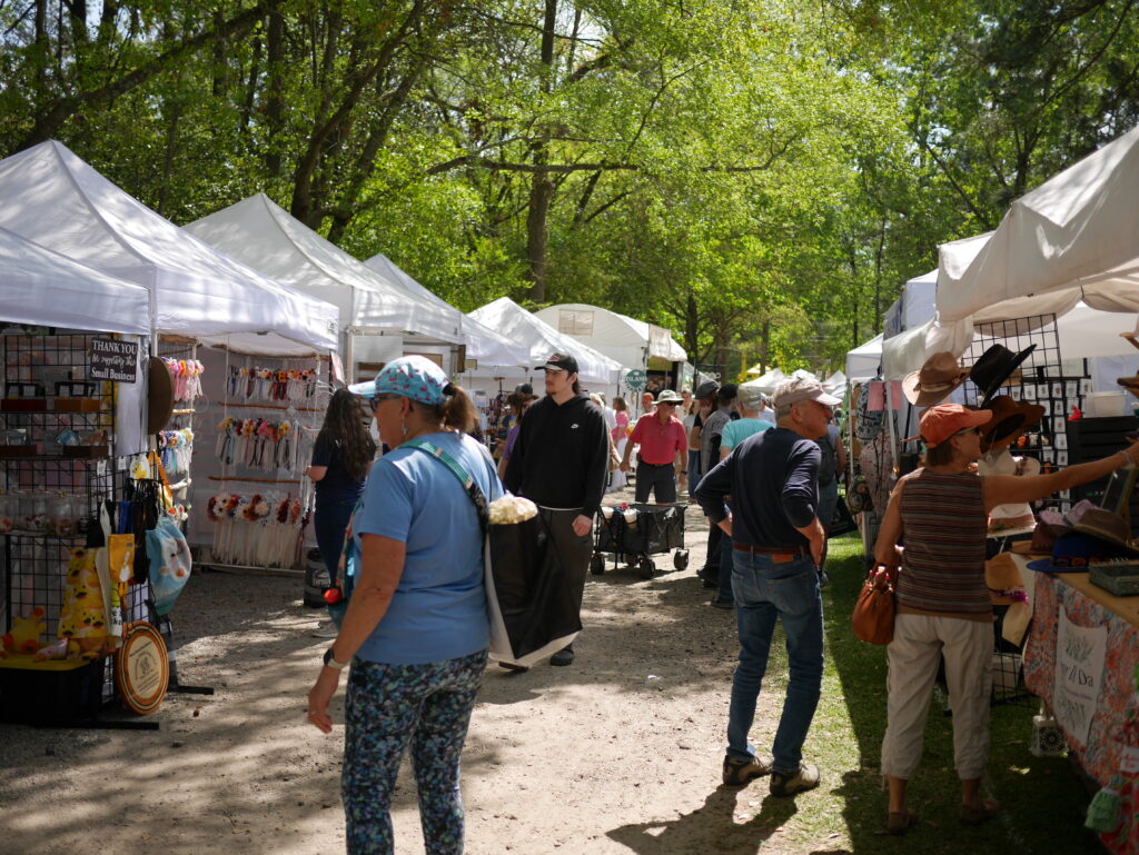 Visitors browse vendor booths at an outdoor market surrounded by trees.