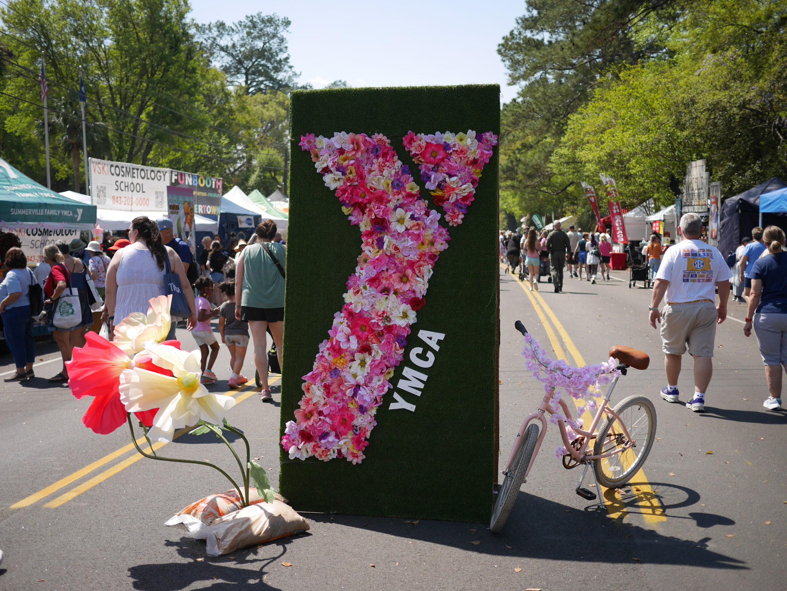 A floral YMCA sign stands beside a decorated bicycle on a busy festival street. People walk in the background.