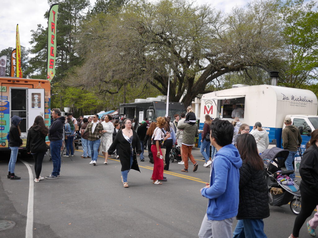 Crowd of people walking among food trucks under trees, some with strollers, enjoying a lively outdoor event.