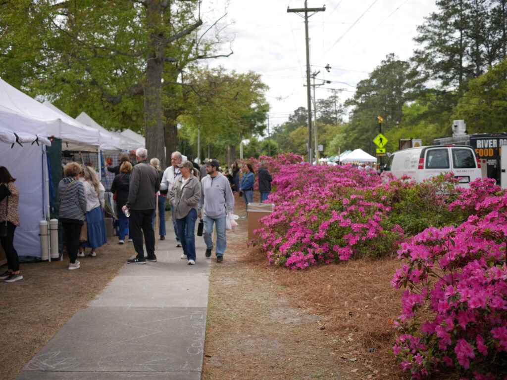 Crowd walks along a path lined with pink azaleas and vendor tents at an outdoor event.
