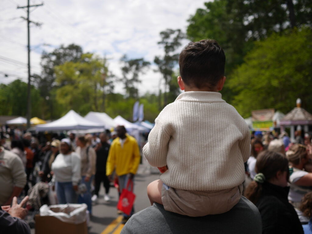 A child sits on an adult's shoulders, overlooking a busy outdoor market with many people and tents.