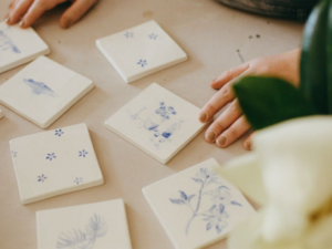 Hands arrange decorative blue and white tiles on a table, surrounded by a plant.