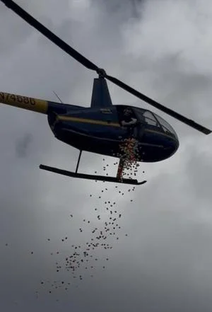A helicopter hovers above, dropping colorful objects from its open door against a cloudy sky.