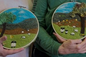 Two people hold embroidered hoop art featuring sheep and landscapes.