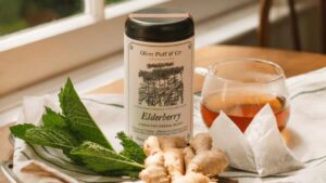 A canister of elderberry tea, fresh ginger, mint leaves, and a glass of brewed tea on a table.