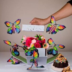 A hand places a card into a colorful pop-up box with flowers and flying butterfly decorations.