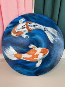 Two koi fish swim in swirling blue water, painted on a round canvas. Background features pink and green fabric.