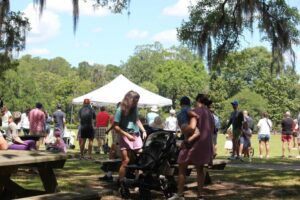 People gather at a park event with a tent; a woman attends to a stroller while others watch from a distance.