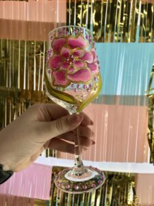 A hand holds a decorated glass with a pink flower design and colorful gems against a shiny backdrop.