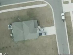 Aerial view of a house with two cars parked in the driveway, surrounded by grass and roads.