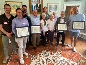 A group of people holds certificates, smiling in a room with framed portraits and a patterned rug.