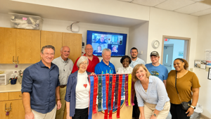 A group of nine people poses with colorful ribbons in front of a video screen displaying remote participants.