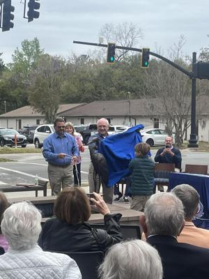 A group of people unveil a blue cloth at an outdoor event, with an audience watching.