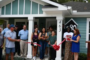A group of people gathers outside a newly built home, preparing to cut a red ribbon.