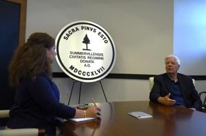 A woman interviews an older man at a table, with a large emblem in the background.