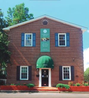 Brick building with green awning, blue shutters, and signage. Flower beds line the entrance.