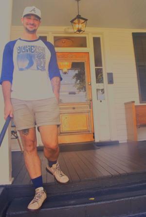 A man stands on a porch, wearing a baseball shirt and shorts, smiling at the camera.