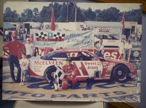 A race car with "McElveen" and "Sweet Jenny" decals is parked. A driver in a red suit poses with fans, including children.