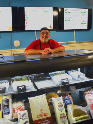 A smiling man in a red shirt stands behind a display case filled with various food items.