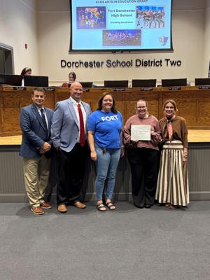 Five people stand in a school board meeting, holding a certificate. A presentation is displayed in the background.