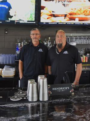 Two bartenders stand behind a bar, smiling. Stainless steel cocktail shakers and a gin display are visible.