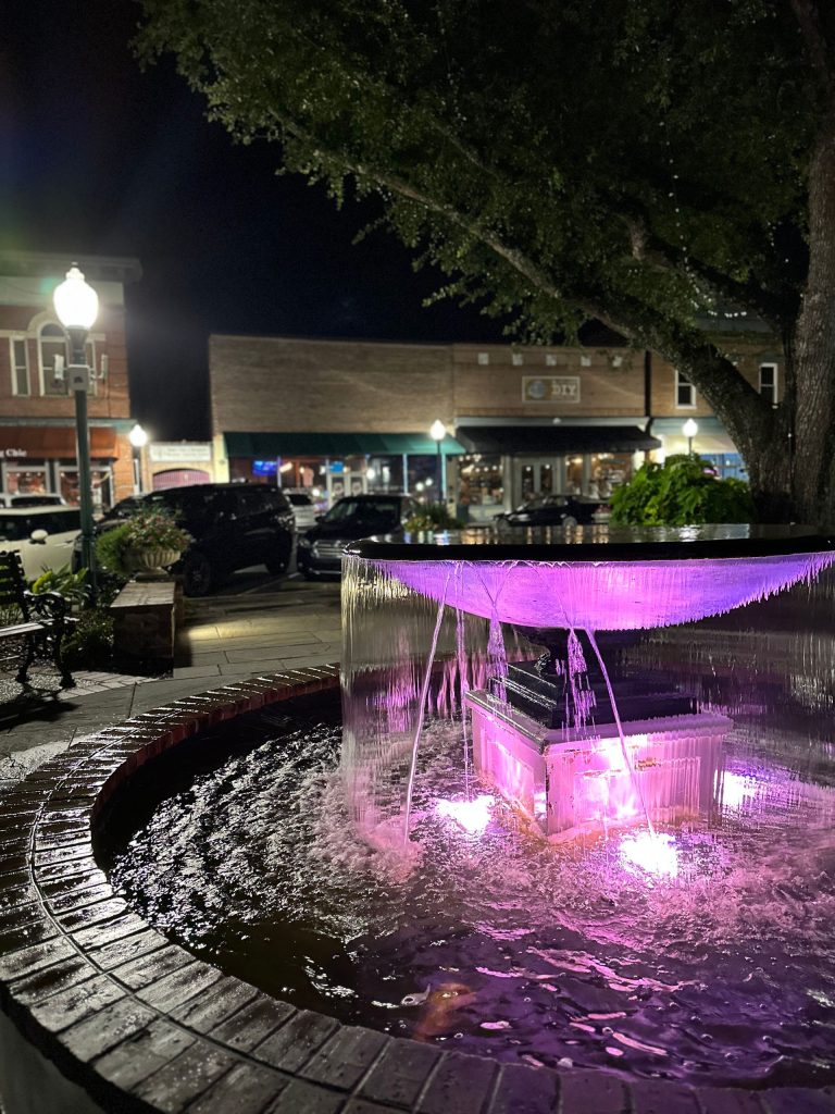 A lit fountain with purple lights flows in a brick-lined basin at night, surrounded by shops and trees.