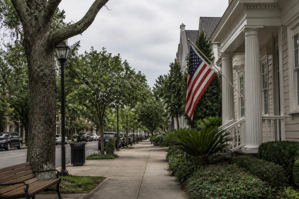 Tree-lined sidewalk with a U.S. flag, benches, and parked cars under a cloudy sky in Summerville, SC.